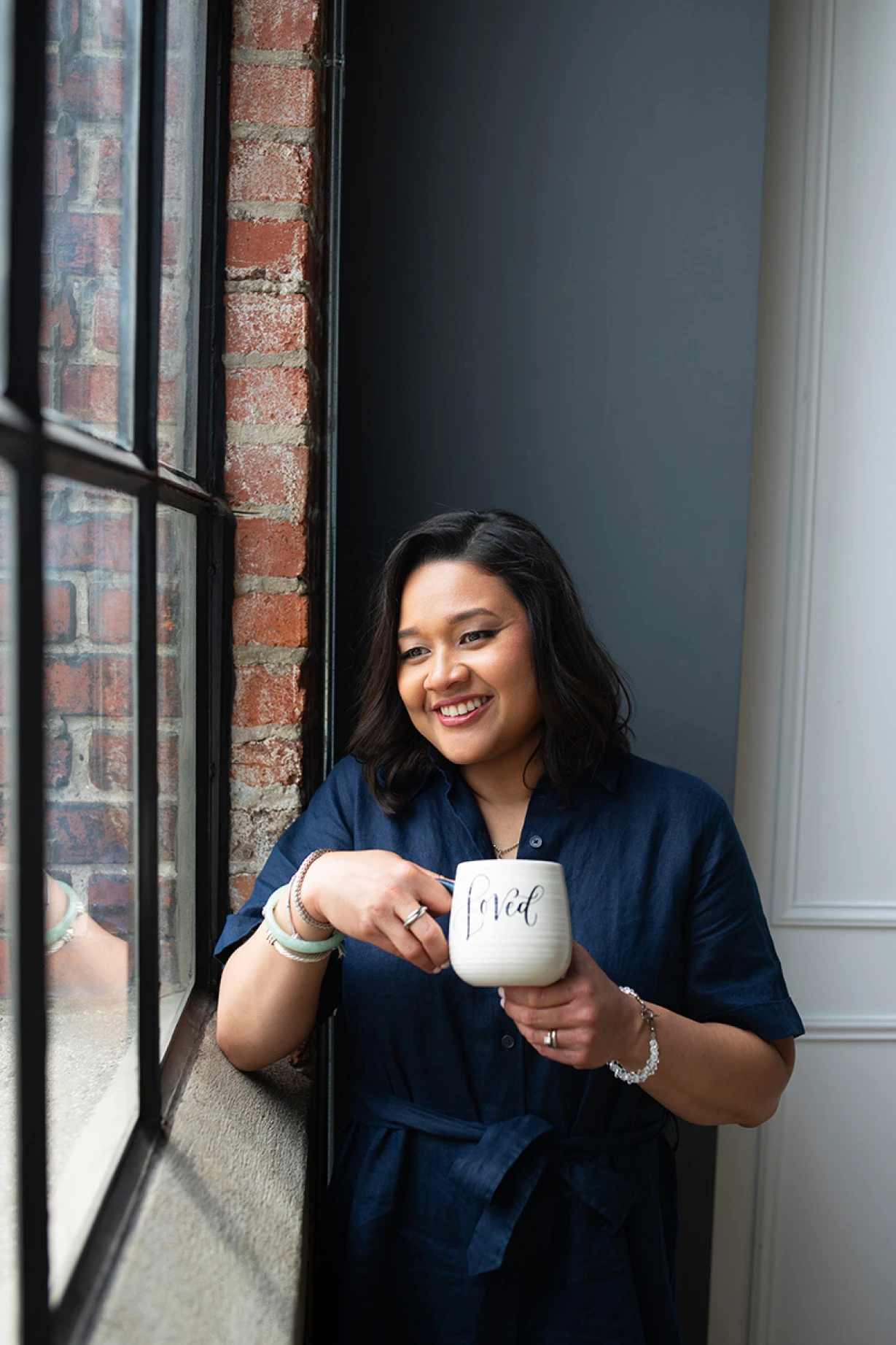 A smiling therapist holding a mug and looking out a window in a relaxed, natural moment